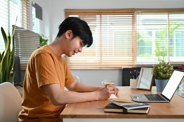 Side view smiling man sitting in front of laptop computer and making notes on notebook