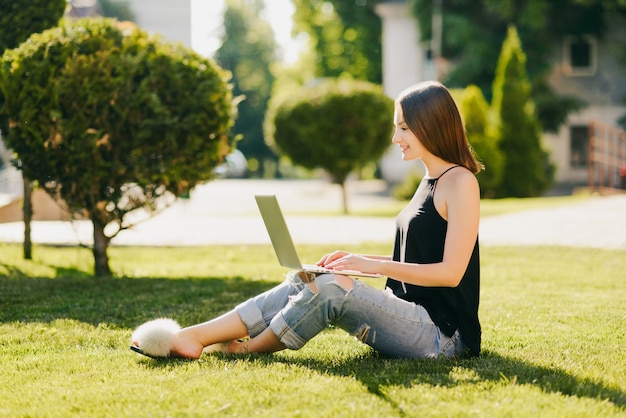 Side view of smiling pretty girl in fashionable clothes sitting on grass and using a laptop computer. In the park.