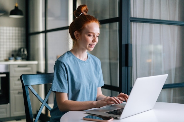 Side view of smiling redhead young woman working typing on laptop computer sitting at table