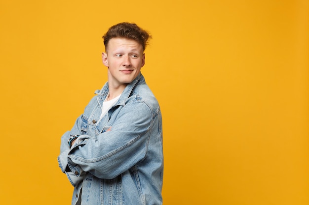 Side view of smiling young man in denim casual clothes holding hands crossed looking back isolated on yellow orange background in studio. People sincere emotions lifestyle concept. Mock up copy space.