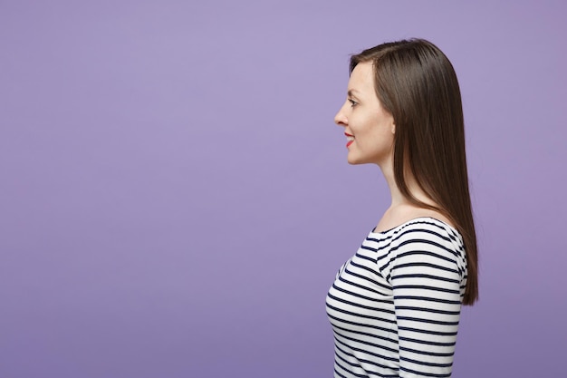 Side view of smiling young woman girl in casual striped clothes posing isolated on violet purple background studio portrait. People lifestyle concept. Mock up copy space. Standing and looking aside.