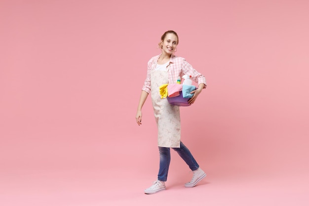 Side view of smiling young woman housewife in apron hold basin with detergent bottles washing cleansers doing housework isolated on pink background in studio. Housekeeping concept. Looking camera.