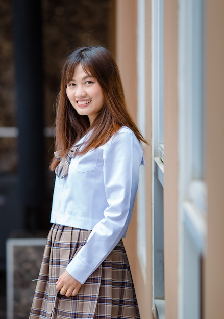 Side view of stylish Asian female student in uniform standing near building of university and looking at camera