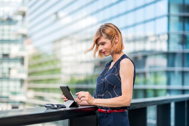 Side view of stylish young businesswoman in classy outfit using tablet while standing near railing against contemporary city building with glass walls