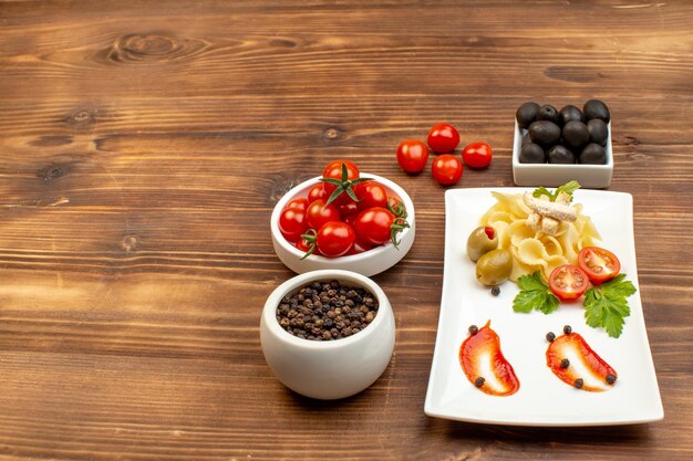 Side view of tasty pasta meal served with vegetables on a white plate tomatoes olives pepper on brown wooden surface