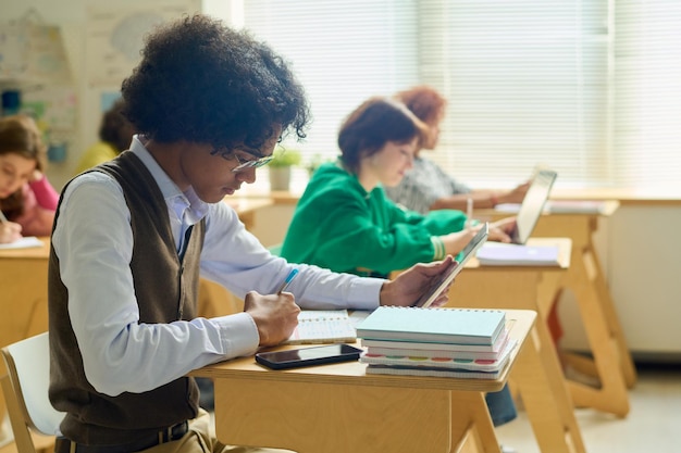 Side view of teenage guy with tablet carrying out individual assignment