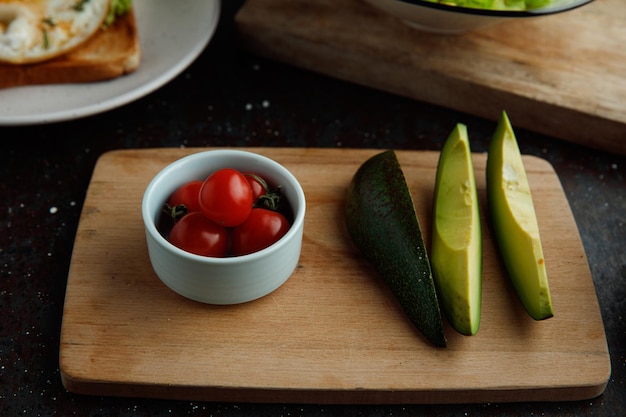 Side view of tomatoes in bowl and sliced avocado on cutting board on black background