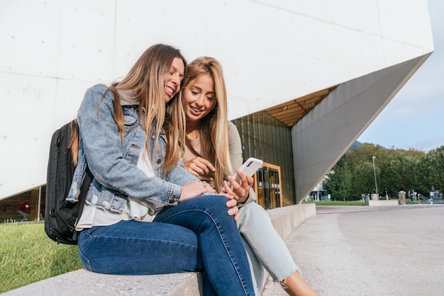 Side view of two female college classmates sitting near of university using phone and having fun.