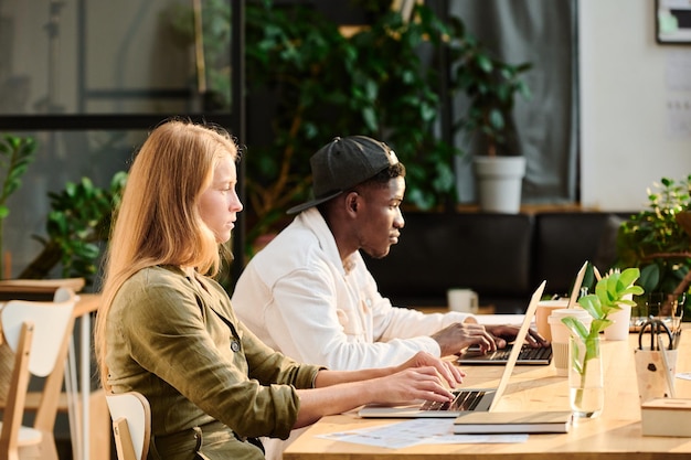 Side view of two young intercultural designers or analysts networking by desk