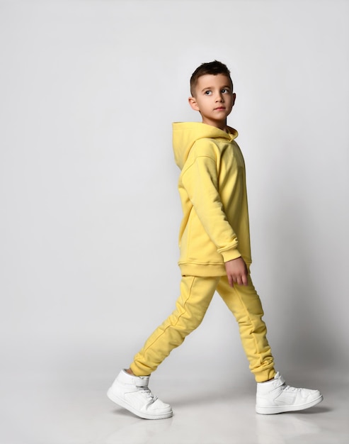 Side view of a walking boy in a yellow tracksuit with a hood and trousers white sneakers isolated on white Portrait of a child in cool clothes Kids fashion casual wear comfortable kids wear