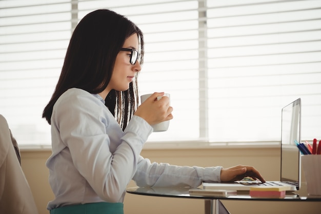 Side view of woman drinking coffee while sitting by laptop