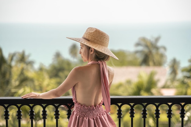 Side view of woman in pink dress and straw hat standing on a hotel balcony, ocean view.