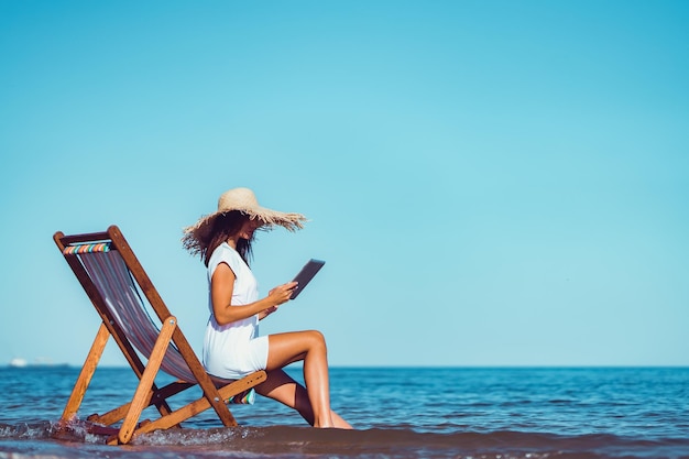 Side view of woman in sunhat who using laptop while lying on the beach chaise longue at the seaside