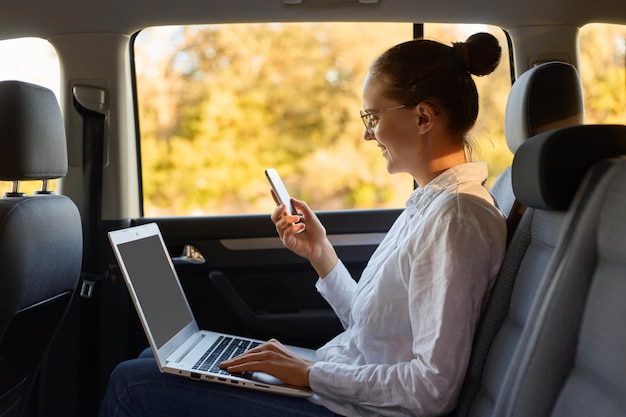 Side view of young adult adorable businesswoman with bun hairstyle wearing white shirt using laptop and mobile phone while sitting in the car going to office and browsing internet