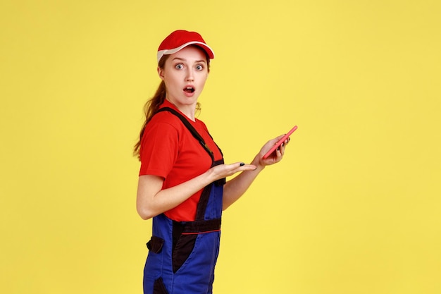 Side view of young adult shocked worker woman using mobile phone for online supporting, expressing astonishment, looking at camera. Indoor studio shot isolated on yellow background.