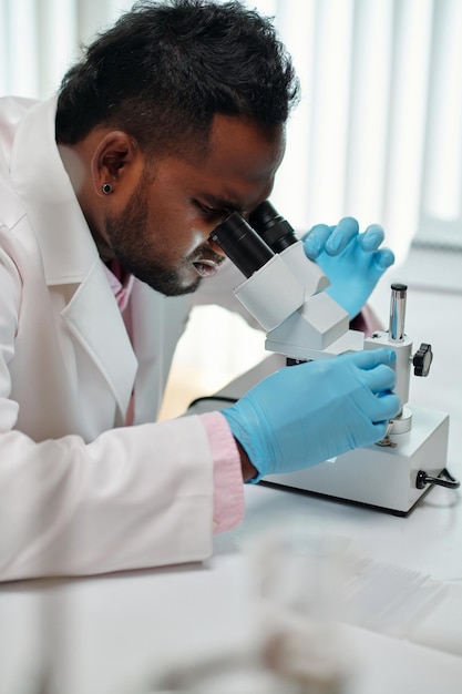 Side view of young african american male scientist looking in microscope while sitting in laboratory