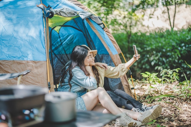 Side view Young Asian pretty woman and her girlfriend sitting at front of tent use mobile phone take photo during camping in forest with happiness together