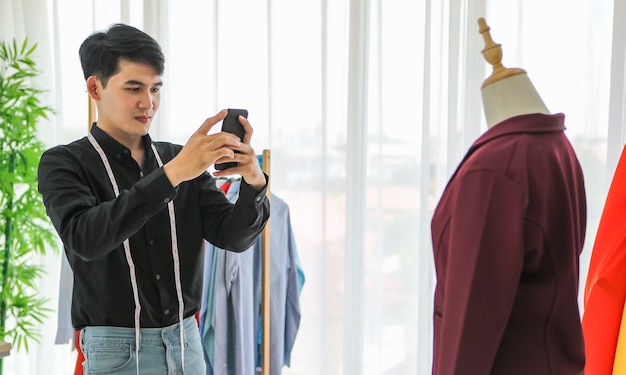 Side view of young Asian tailor using smartphone to shoot trendy jacket on dummy for portfolio.