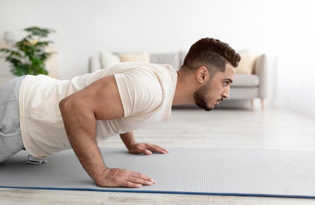 Side view of young athletic arab man doing pushups on sports mat at home copy space