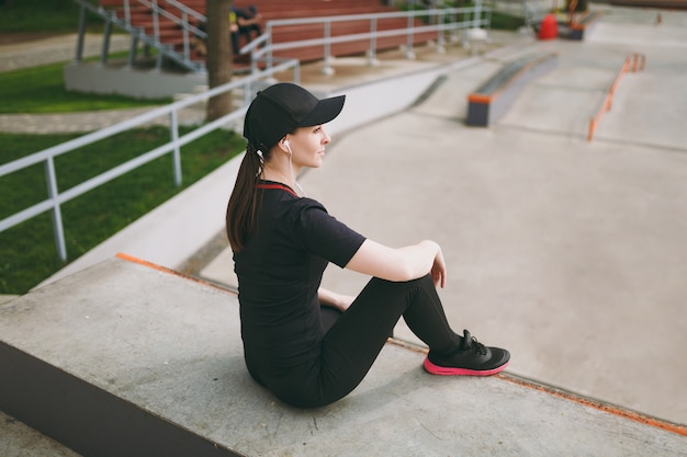 Photo side view young athletic brunette woman in black uniform and cap with headphones listening to music resting and sitting before or after running, training in city park outdoors