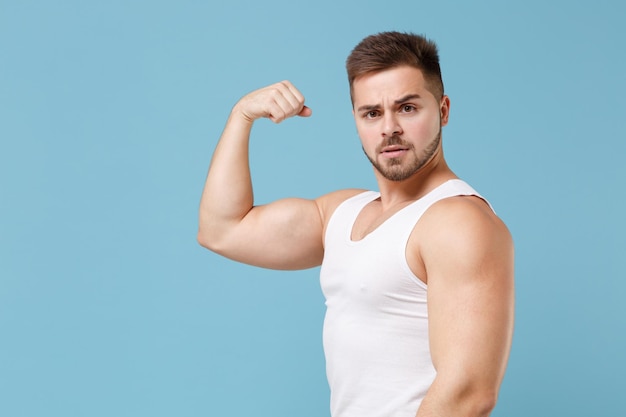 Side view of young bearded guy 20s in white singlet posing isolated on pastel blue wall background studio portrait. Sport fitness healthy lifestyle concept. Mock up copy space. Showing biceps muscles.