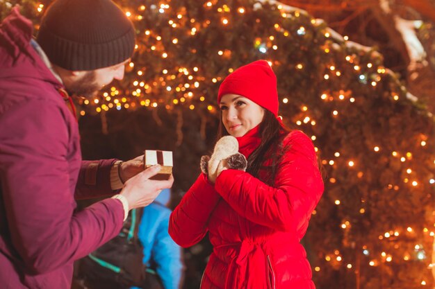 Side view of young bearded man making proposal of marriage on Christmas eve outdoors to his cute brunette lady, she is happy and amazed. Lights on Christmas arch on background