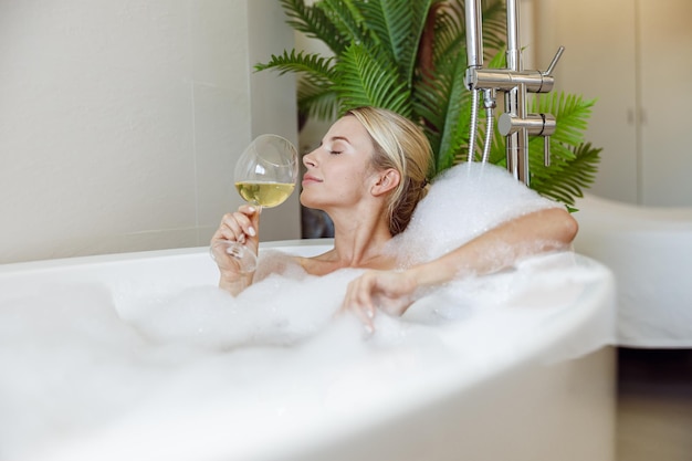 Side view on young beautiful caucasian female enjoying white wine and taking bath bathroom