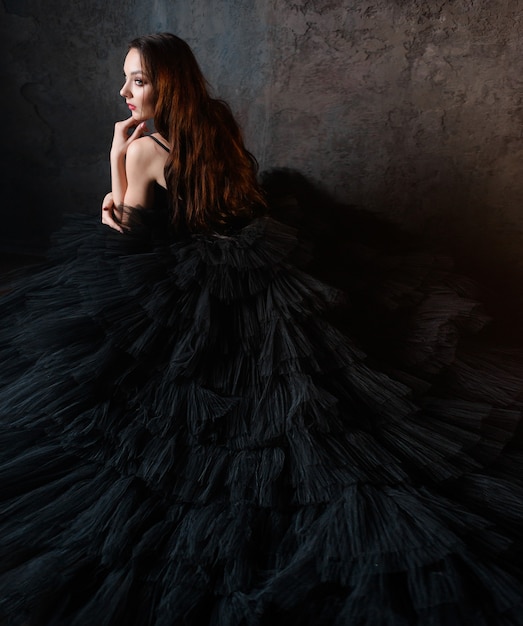 Side view of young brunette in a lush black dress sitting near the wall