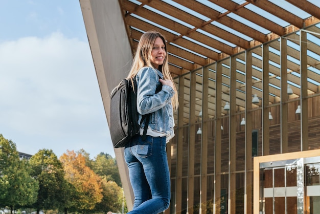 Side view of a young college girl holding her backpack on campus looking at the camera.