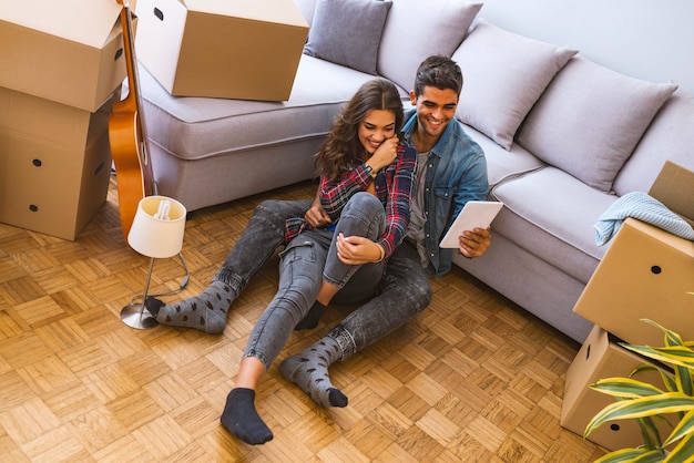 Side view of young couple sitting on floor near carton boxes and browsing modern laptop while moving into new apartment together 