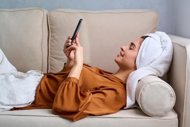 Side view of young happy female with smartphone relaxing on beige sofa