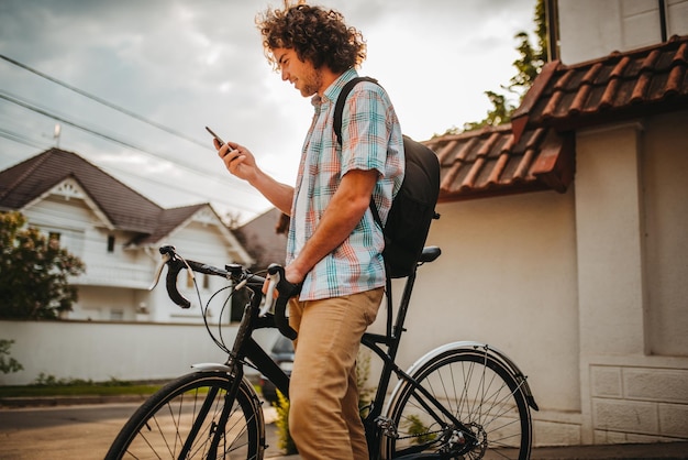 Side view of young happy male with curly hair looking on his mobile phone choosing gps application for the road for bicycle Happy smiling guy cyclist with backpack surfing internet using smart phone