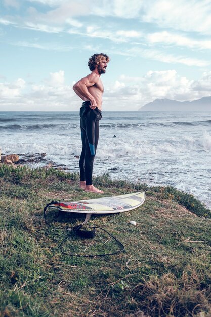 Side view of young muscular Hispanic male surfer putting on wetsuit while standing on grassy coast near board before practicing surfing in wavy ocean