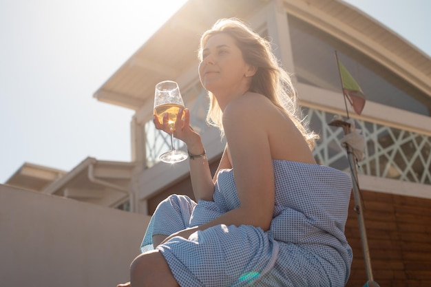 Side view of young sensual woman with fair hair wrapped in blue sheet holding glass of wine sitting