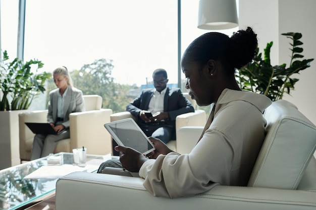 Side view of young serious female office worker with tablet sitting on couch