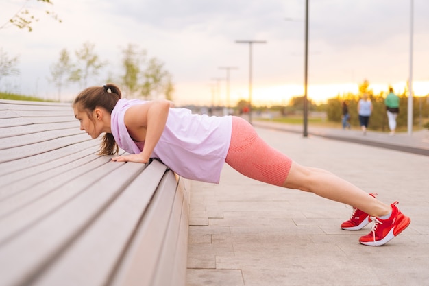 Side view of young sportive woman wearing activewear training doing push ups using street bench in