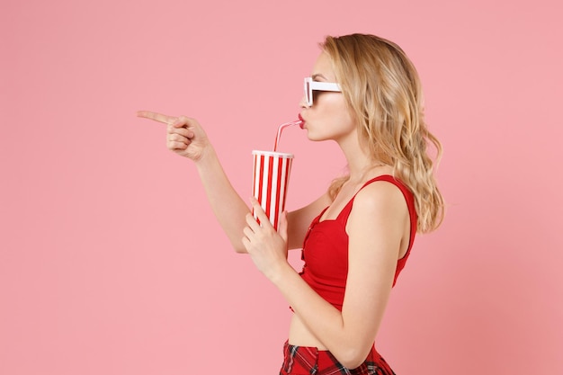 Side view of young woman in red sexy clothes, dark sunglasses isolated on pastel pink background. People lifestyle concept. Mock up copy space. Hold cup of cola or soda, pointing index finger aside.