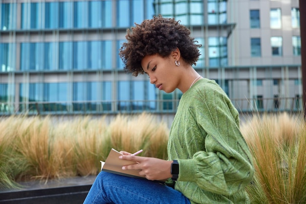 Sideways shot of attentive curly haired young woman makes notes in notepad with pen makes creative task wears casual green jumper and smartwatch sits outdoors near city buildings works in open air.