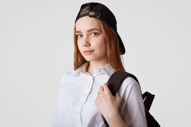 Sideways shot of beautiful schoolgirl wears trendy black cap, white shirt, carries backpack, ready for school and lessons, isolated over white wall School, studying and education concept