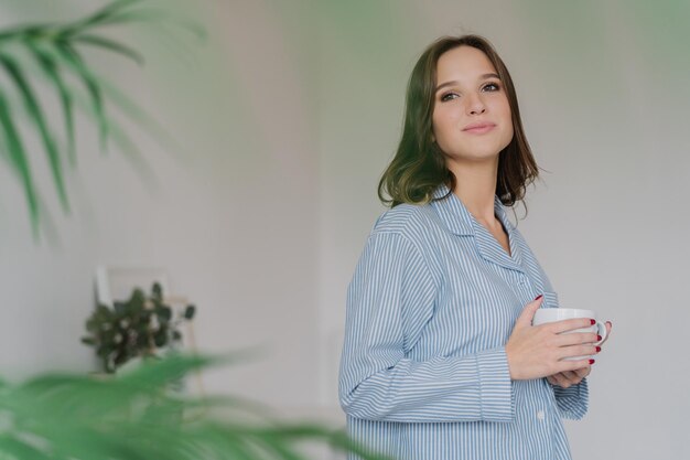 Sideways shot of dreamy Caucasian woman dressed in casual clothes drinks tea stands indoor against domestic interior with copy space for your advertising content People rest and drinking concept