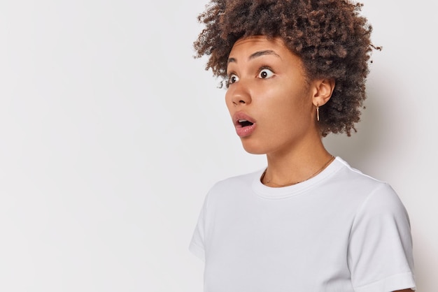 Sideways shot of shocked impressed woman stares bugged eyes keeps mouth widely opened notices horrible scene into distance weaars casual basic t shirt isolated over white background blank space