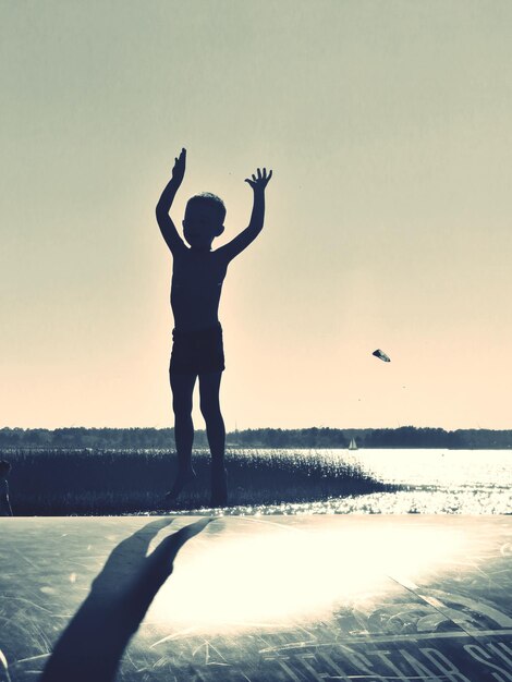 Photo silhouette boy jumping by lake and clear sky during sunset