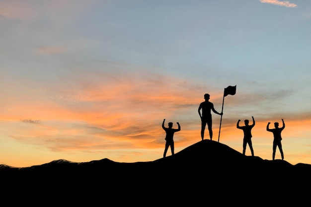 Silhouette of business leader holding a flag and employee making high hands over head on top of the mountain in beautiful sunset sky evening time for business success and teamwork concept in company