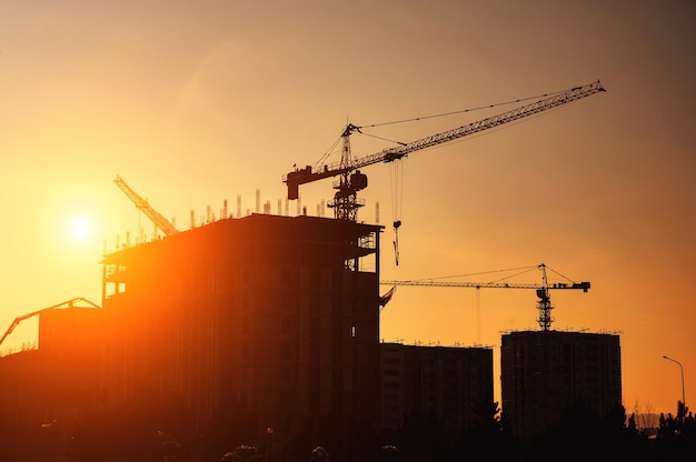 Silhouette of construction crane building a house on a construction site against background of sunset sky