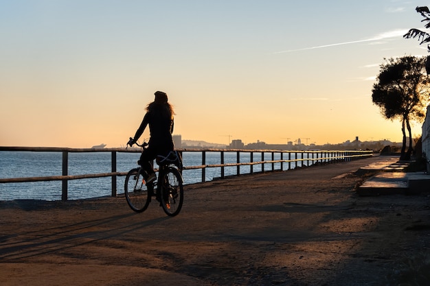 Silhouette of curly woman riding a bicycle at the sunset on the promenade road