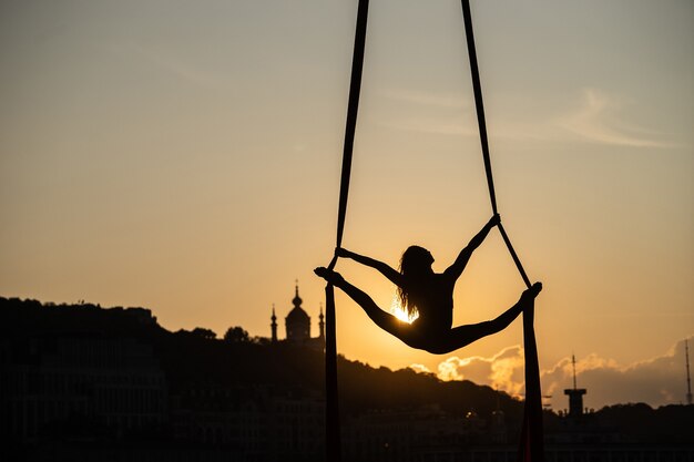 Silhouette of a flexible woman acrobat on aerial silk during a sunset on Kiev city. concept of freedom and peace.