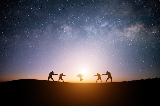 Silhouette of four young businessman pulling tug of war with a rope on top of the mountain with night sky and star background Concept for business competition rivalry challenge or dispute