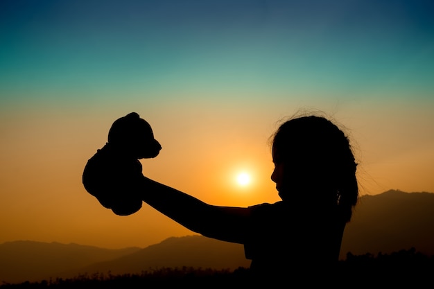 silhouette of girl standing alone and hand holding teddy bear at a sunset time