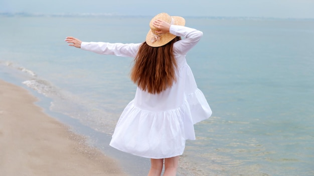 Silhouette of happy enjoying woman from back with thin legs in white beach dress having rest at sunset on sea beach Unrecognizable happy woman in straw hat in hand on seashore Long web banner