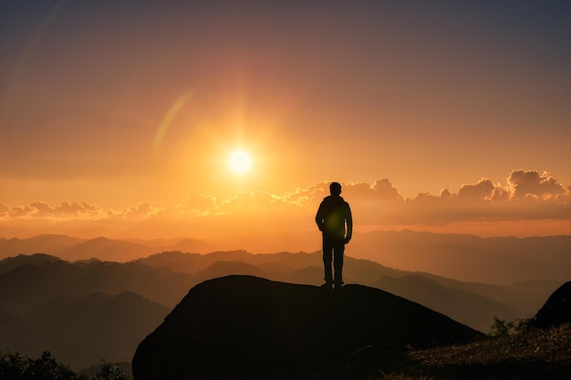 Silhouette hiker man standing on top of mountain with the sun shining over mountain in the sunset